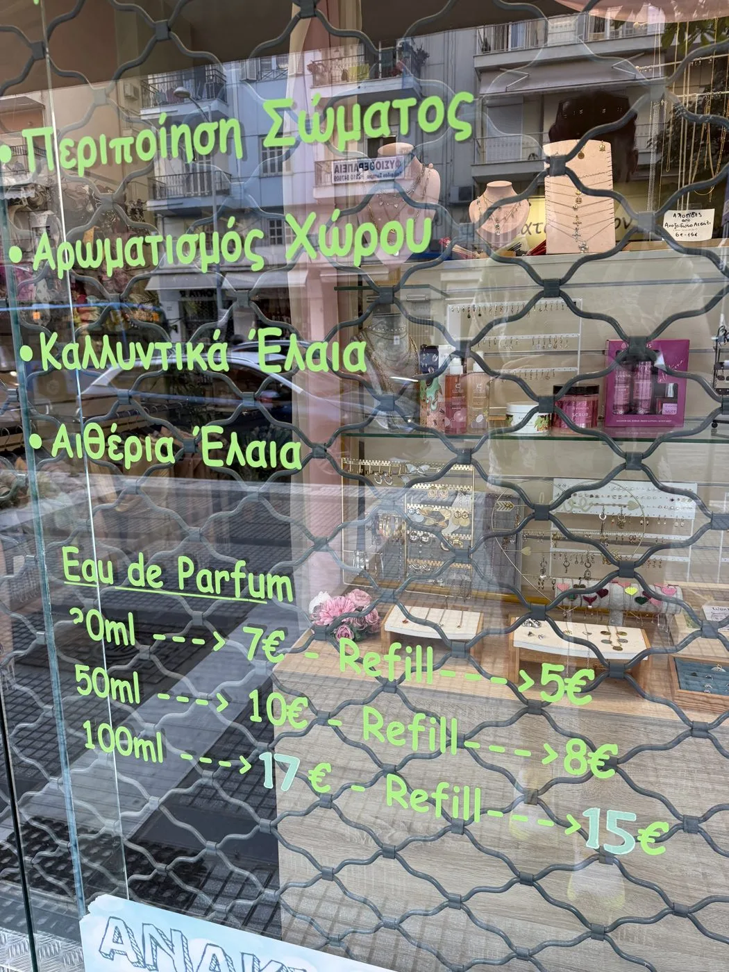 The shop window in Kalamaria: a hand-lettered green price list on the glass, the security grille drawn behind it, the 8 being the refill price.